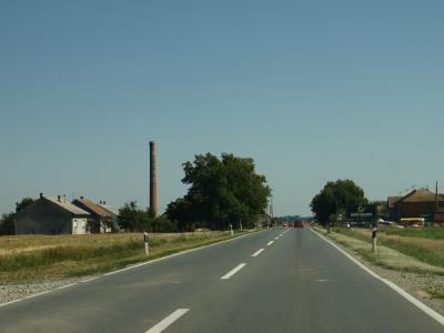 A paved two-lane road stretching through a rural landscape with scattered houses, trees, and a tall brick chimney on the left; a billboard for 'RADIO BARANJA' is visible on the right side of the road.