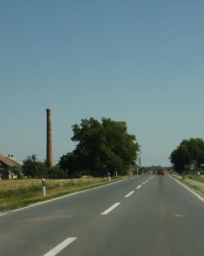 A paved two-lane road stretching through a rural landscape with scattered houses, trees, and a tall brick chimney on the left; a billboard for 'RADIO BARANJA' is visible on the right side of the road.