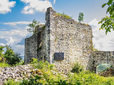 A weathered stone ruin of a medieval fortress wall and tower, featuring a mounted commemorative plaque and an arched opening, set against a backdrop of green hills and a blue sky.