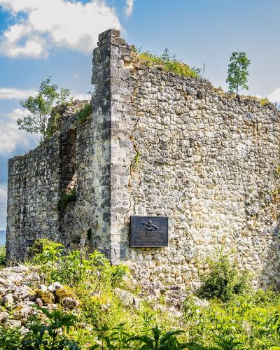 A weathered stone ruin of a medieval fortress wall and tower, featuring a mounted commemorative plaque and an arched opening, set against a backdrop of green hills and a blue sky.