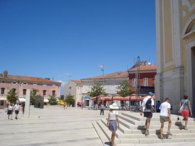A wide, paved public square with people walking and sitting at outdoor cafes, featuring a large yellow church facade on the right and pinkish buildings in the background under a clear blue sky.