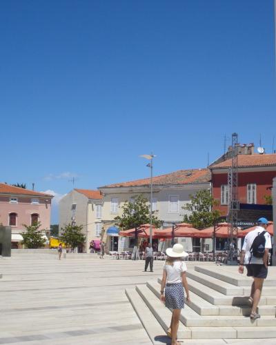 A wide, paved public square with people walking and sitting at outdoor cafes, featuring a large yellow church facade on the right and pinkish buildings in the background under a clear blue sky.