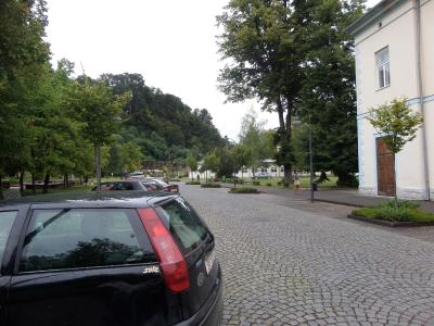 A black Volvo station wagon is parked on a cobblestone street in the foreground, with a cream-colored building featuring blue trim and a wooden door visible on the right, and a park-like area with trees and other buildings in the background.