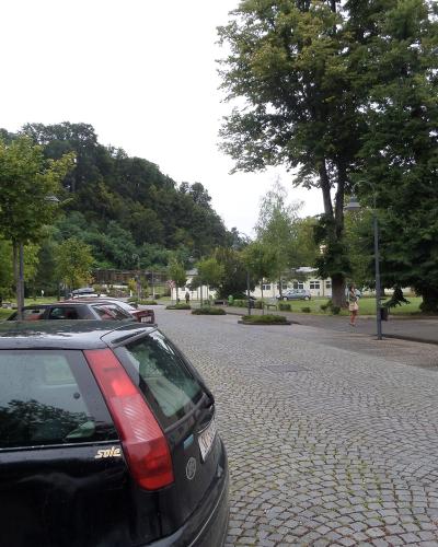 A black Volvo station wagon is parked on a cobblestone street in the foreground, with a cream-colored building featuring blue trim and a wooden door visible on the right, and a park-like area with trees and other buildings in the background.