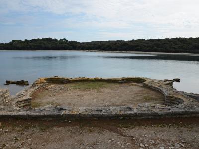 The image shows the stone foundations of an ancient rectangular structure with curved corners situated directly on the shoreline of a calm bay, with a forested island coast in the background.