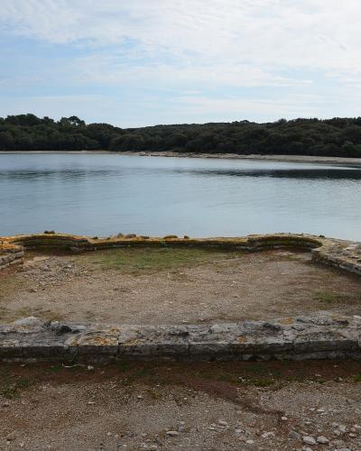 The image shows the stone foundations of an ancient rectangular structure with curved corners situated directly on the shoreline of a calm bay, with a forested island coast in the background.