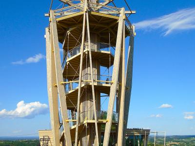 A tall, modern wooden observation tower with a spiral staircase and viewing platforms, set on a grassy hilltop under a blue sky with rolling green countryside in the background.