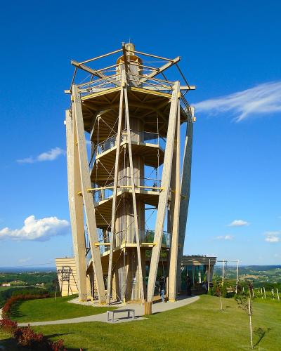 A tall, modern wooden observation tower with a spiral staircase and viewing platforms, set on a grassy hilltop under a blue sky with rolling green countryside in the background.
