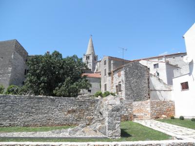 A view of the historic stone town of Bale in Croatia, featuring old stone buildings, a prominent church tower with a conical roof, and a stone wall in the foreground.