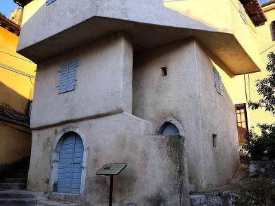 A two-story, off-white stucco building with a distinctive overhanging upper floor, blue louvered shutters, and an arched doorway, situated on a cobblestone street with an informational plaque in front.