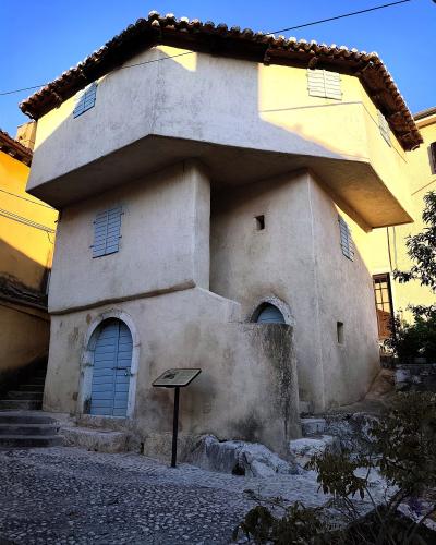 A two-story, off-white stucco building with a distinctive overhanging upper floor, blue louvered shutters, and an arched doorway, situated on a cobblestone street with an informational plaque in front.