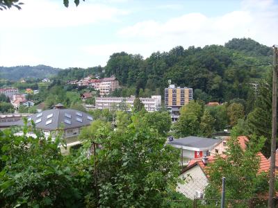 A scenic view of a town nestled in a green valley with several multi-story buildings, including a prominent yellow and white hotel-like structure and a building with a distinctive dark, sloped roof with skylights, surrounded by trees and hills.