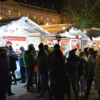A night scene of a crowded outdoor Christmas market with white wooden stalls, string lights, and people in winter clothing; one stall sign reads "ZAGORSKI ŠTRUKLI" and another says "Bor Bor".