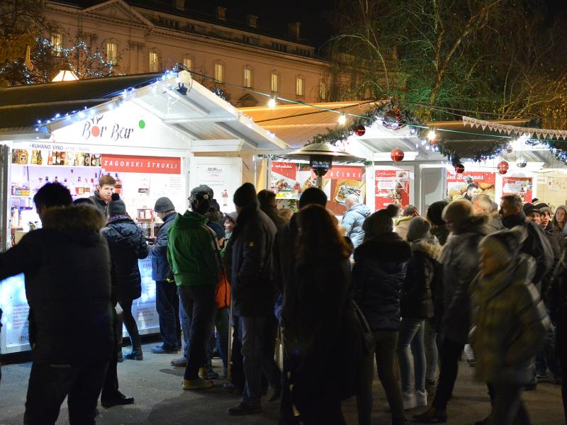 A night scene of a crowded outdoor Christmas market with white wooden stalls, string lights, and people in winter clothing; one stall sign reads "ZAGORSKI ŠTRUKLI" and another says "Bor Bor".
