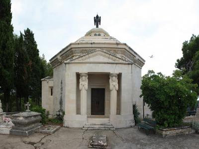 A hexagonal stone mausoleum featuring two caryatid columns at the entrance and a dark bird sculpture on the roof, situated within a cemetery surrounded by trees.