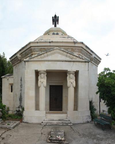 A hexagonal stone mausoleum featuring two caryatid columns at the entrance and a dark bird sculpture on the roof, situated within a cemetery surrounded by trees.