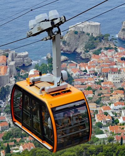 A high-angle view from a cable car looking down at the walled Old Town of Dubrovnik, Croatia, with the cable car support tower and cables visible in the foreground.