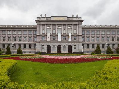 A large, grey, multi-story neo-Renaissance building with the name "MIMARA" in gold letters on the central pediment, fronted by a manicured lawn and a large flower bed.