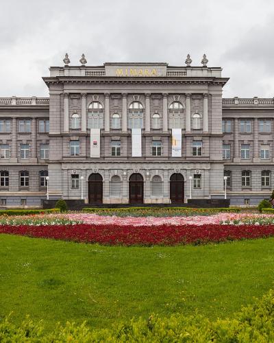 A large, grey, multi-story neo-Renaissance building with the name "MIMARA" in gold letters on the central pediment, fronted by a manicured lawn and a large flower bed.