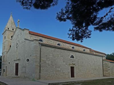 A stone church with a tall, slender bell tower on the left, a terracotta tiled roof, and a large pine tree branch overhanging the top right, set against a blue sky.