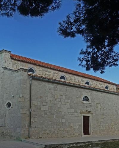 A stone church with a tall, slender bell tower on the left, a terracotta tiled roof, and a large pine tree branch overhanging the top right, set against a blue sky.