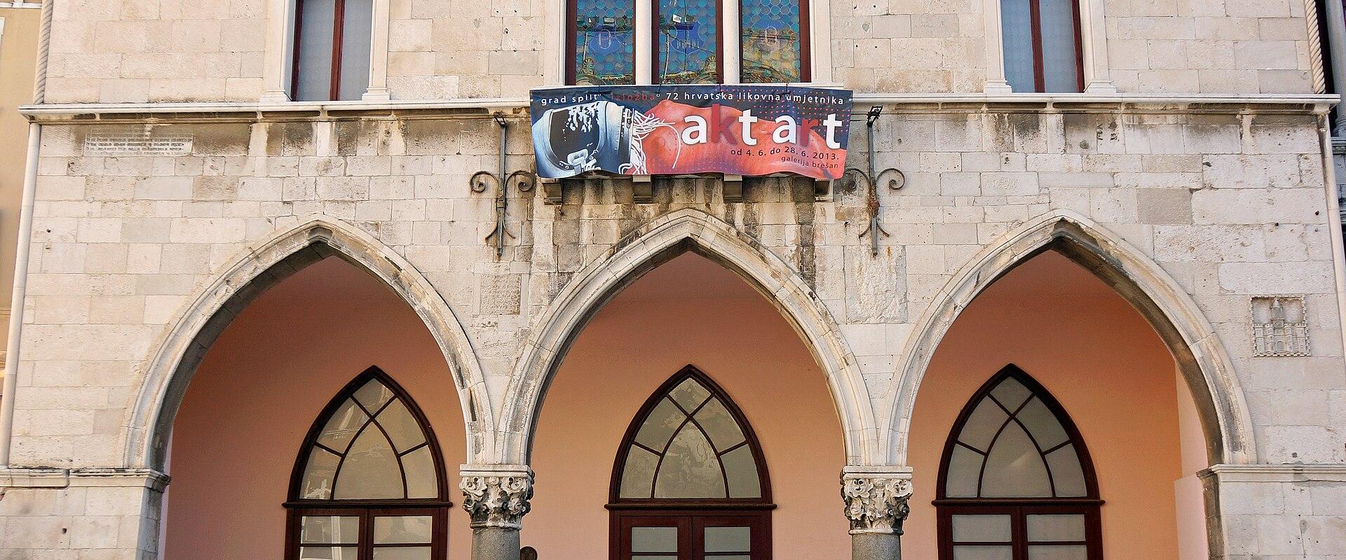 A three-story stone building with Gothic-style arched windows and a ground-floor loggia featuring three large pointed arches supported by columns, with a banner reading "grad split" and "aktat" hanging above the central arch.