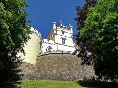 A low-angle view of a white castle with crenellated towers and a stone retaining wall, framed by green trees under a blue sky, with two Croatian flags flying from the roof.
