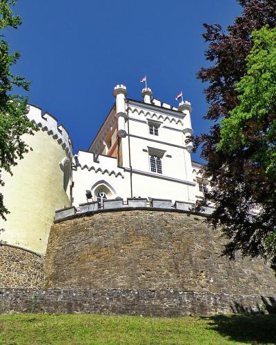 A low-angle view of a white castle with crenellated towers and a stone retaining wall, framed by green trees under a blue sky, with two Croatian flags flying from the roof.