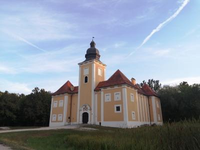 A yellow Baroque-style building with a central tower topped by a dark dome and red-tiled roofs, set against a blue sky with contrails and surrounded by greenery.