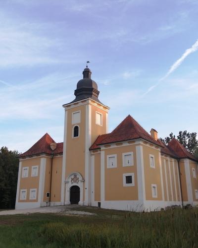 A yellow Baroque-style building with a central tower topped by a dark dome and red-tiled roofs, set against a blue sky with contrails and surrounded by greenery.