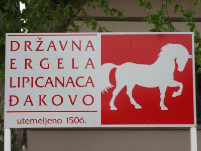 A rectangular sign with a red background on the right featuring a white silhouette of a horse, and text on the left reading "DRŽAVNA ERGELA LIPICANACA ĐAKOVO" and "utemeljeno 1506."