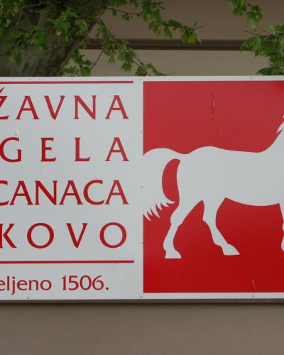 A rectangular sign with a red background on the right featuring a white silhouette of a horse, and text on the left reading "DRŽAVNA ERGELA LIPICANACA ĐAKOVO" and "utemeljeno 1506."
