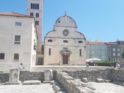 The image shows a large stone church with a prominent bell tower and a distinctive facade featuring a rose window and a curved pediment, situated in an urban square with ancient Roman ruins in the foreground.