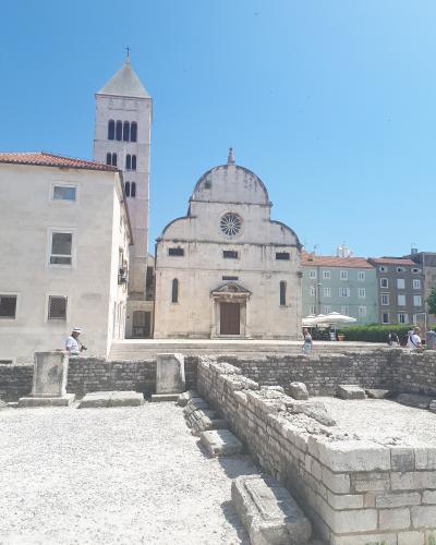 The image shows a large stone church with a prominent bell tower and a distinctive facade featuring a rose window and a curved pediment, situated in an urban square with ancient Roman ruins in the foreground.