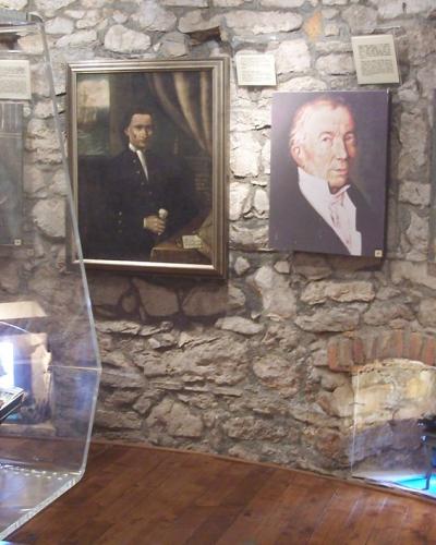 Interior of a museum featuring a large model sailing ship in a glass case, portraits of historical figures on a stone wall, and a display case with documents.