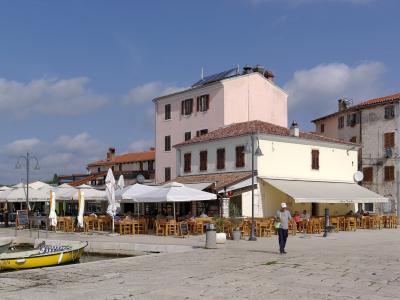 A paved waterfront promenade in a coastal town featuring outdoor restaurant seating under white umbrellas, a yellow boat moored at the edge, and multi-story buildings with terracotta roofs and shuttered windows under a blue sky.