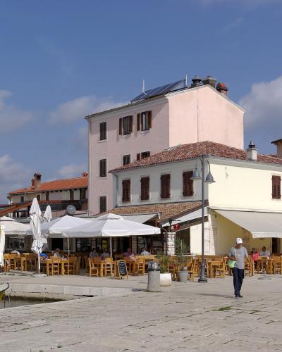 A paved waterfront promenade in a coastal town featuring outdoor restaurant seating under white umbrellas, a yellow boat moored at the edge, and multi-story buildings with terracotta roofs and shuttered windows under a blue sky.