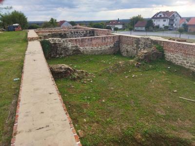 Low stone and brick ruins of a building foundation, a concrete walkway, and a grassy area, with residential houses and a road visible in the background.