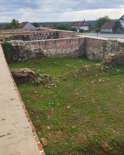 Low stone and brick ruins of a building foundation, a concrete walkway, and a grassy area, with residential houses and a road visible in the background.