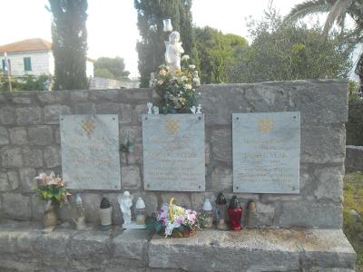 A stone memorial wall featuring three marble plaques with Croatian inscriptions and the national checkerboard coat of arms, adorned with flowers, candles, and small statues.