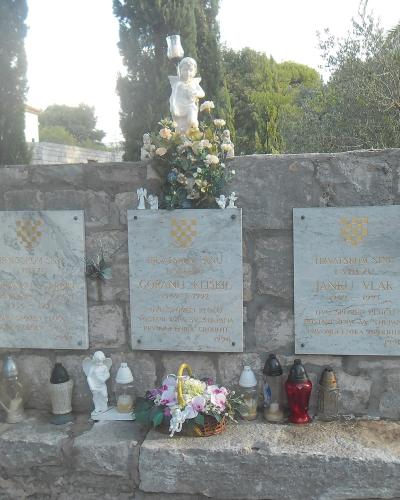 A stone memorial wall featuring three marble plaques with Croatian inscriptions and the national checkerboard coat of arms, adorned with flowers, candles, and small statues.