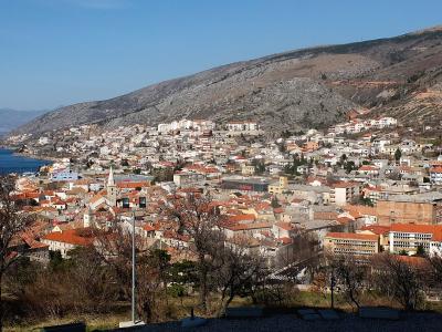 A panoramic view of a coastal town with red-tiled roofs nestled at the foot of a large, rocky hill, overlooking a blue sea under a clear sky.
