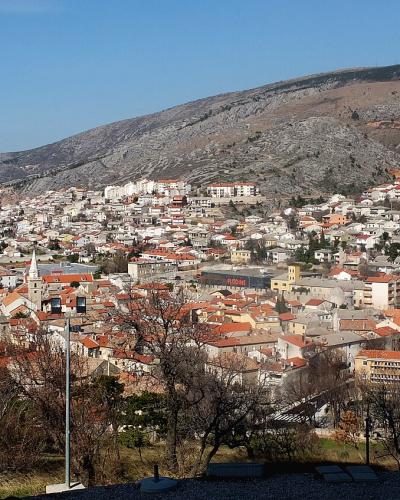 A panoramic view of a coastal town with red-tiled roofs nestled at the foot of a large, rocky hill, overlooking a blue sea under a clear sky.