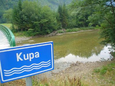 A blue rectangular sign with the white text "Kupa" and three wavy lines indicating a river, positioned in the foreground next to a green bridge railing overlooking a flowing river and forested banks.