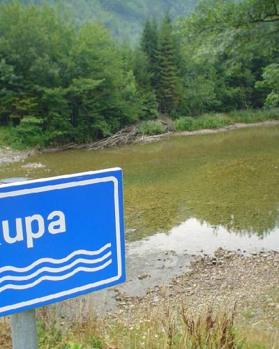 A blue rectangular sign with the white text "Kupa" and three wavy lines indicating a river, positioned in the foreground next to a green bridge railing overlooking a flowing river and forested banks.