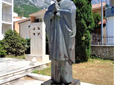 A bronze statue of a bishop in prayer standing on a stone pedestal with the inscription "BLAŽENI ALOJZIJE STEPINAC 1898 - 1960", set outdoors with mountains in the background.