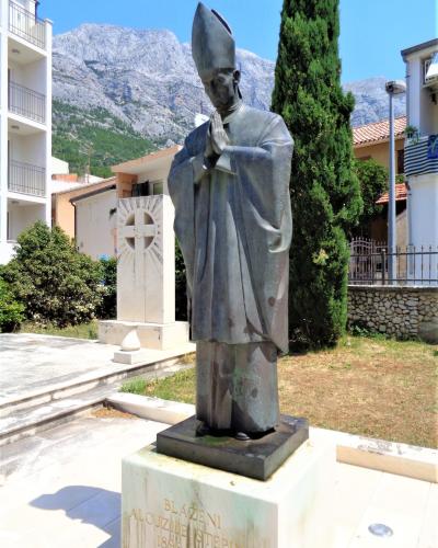 A bronze statue of a bishop in prayer standing on a stone pedestal with the inscription "BLAŽENI ALOJZIJE STEPINAC 1898 - 1960", set outdoors with mountains in the background.
