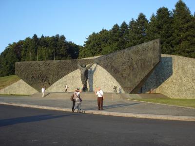A large, modernist bronze and stone monument featuring a central figure with raised arms flanked by relief panels, situated in an open paved plaza with trees in the background.
