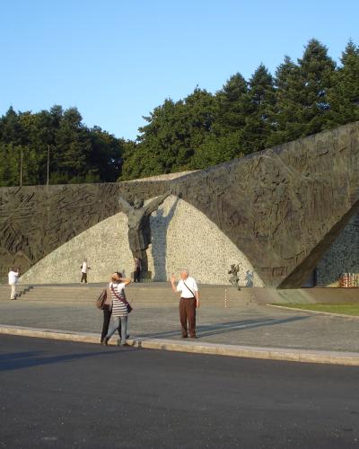 A large, modernist bronze and stone monument featuring a central figure with raised arms flanked by relief panels, situated in an open paved plaza with trees in the background.