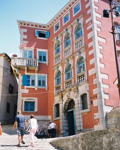 A tall, salmon-pink building with white quoins and blue shutters, featuring a grand stone staircase leading up to an arched entrance, with the word "MUSEUM" carved into the stone facade near the ground floor.